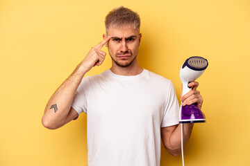 Young caucasian man holding an iron isolated on yellow background showing a disappointment gesture...