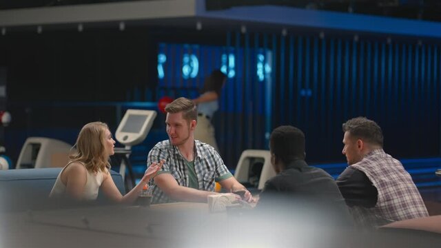 A multi ethnic group of friends in a bowling club are chatting at the table and smiling, rejoicing and high-fiving a friend who knocked out a strike