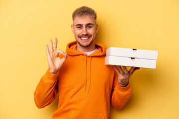 Young caucasian man holding pizzas isolated on yellow background cheerful and confident showing ok gesture.