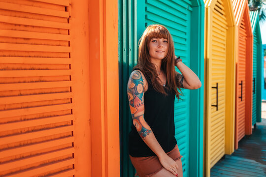Young smiling woman with a tattoo on her arm, standing leaning by beach's changing room. Multicolored wooden cabins on the background