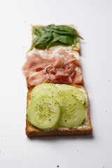 White toasts with cream cheese, ham, cucumbers and spinach on a white surface