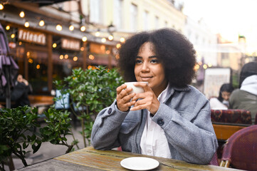 Fashion street style portrait of attractive young natural beauty African American woman with afro hair in tweed jacket posing outdoors in sidewalk cafe. Happy lady with coffee cup in big city