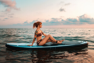 A tanned young woman with a tattoo poses sitting on a sup board swimming at water. Copy space. Sunset sky. The concept of sports recreation and surfing