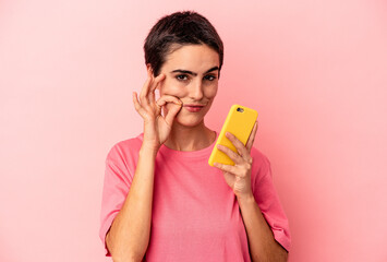 Young caucasian woman holding mobile phone isolated on pink background with fingers on lips keeping...
