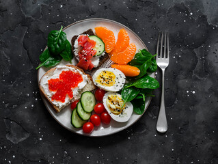 Breakfast, snack, tapas plate - sandwiches with red caviar, salmon, cream cheese, vegetables, spinach, boiled egg on a dark background, top view