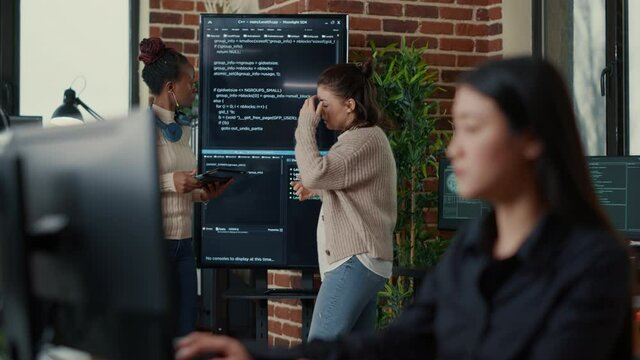 System engineers comparing source code on wall screen tv analyzing errors using digital tablet with focus switching to programer writing algorithm. Team of coders debugging machine learning data.