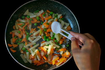 Female hand adding salt to vegetables in pan closeup. Making vegetable isolated on black. Selective...