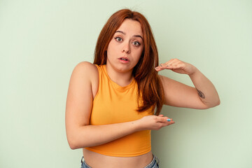 Young caucasian woman isolated on green background holding something with both hands, product presentation.