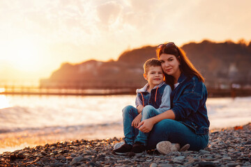 A young mother and her son sitting together and hugs on the beach. In the background is the sea and sunset. Copy space. The concept of a happy childhood