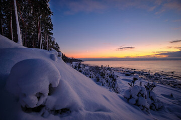 deep snow over rocks at the beach in cold winter sunset by the sea with pine trees and spruce