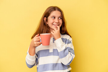 Little caucasian girl holding a pink mug isolated on yellow background relaxed thinking about something looking at a copy space.