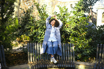 Fashion street style portrait of attractive young natural beauty African American woman with afro hair in blue coat and jeans posing outdoors. Happy tourist laughing walks around the city