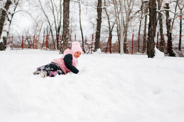Winter portrait of female kid smiling and lying outside in snow in warm winter clothes with high trees around. Adorable background full of snow.