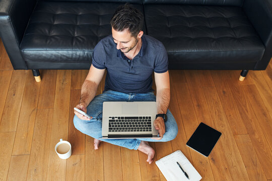 Smiling man working at home using smartphone and laptop sitting on the floor