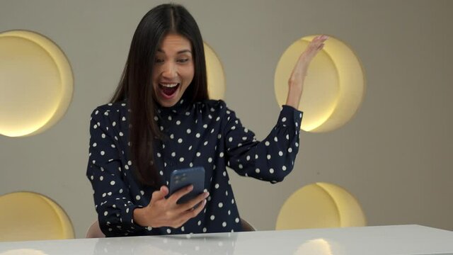 An Asian woman sits at her desk in the office, holds a smartphone, receives a message from the bank and feels incredibly happy. The woman makes a gesture look very excited, winning the lottery