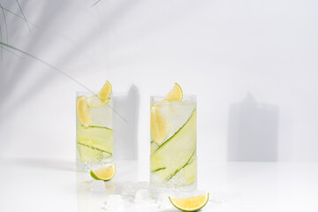 Dos vasos de bebida desintoxicante, agua con fruta y hielo. Contiene pepino, lima y hojas verdes sobre una mesa blanca. Con suave sombra en la pared. Verano, concepto de cóctel tropical y fresco