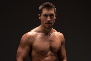 Fototapeta na wymiar Waist up view of the portrait of shirtless bodybuilder looking at the camera while posing over the black wall. Stock photo