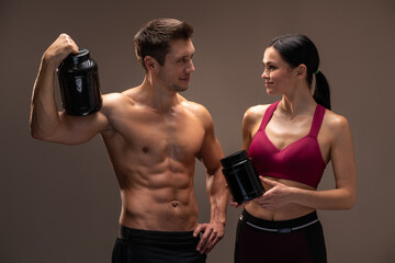 Waist up portrait of sporty beautiful smiling woman in sportswear holding shaker with healthy protein drink and looking at man isolated over brown studio background