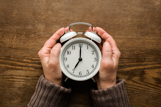 Old mature woman hands holding white alarm clock on dark brown wooden table background. Closeup. Point of view shot. Seven o'clock. Concept of female biological time. Top down view.