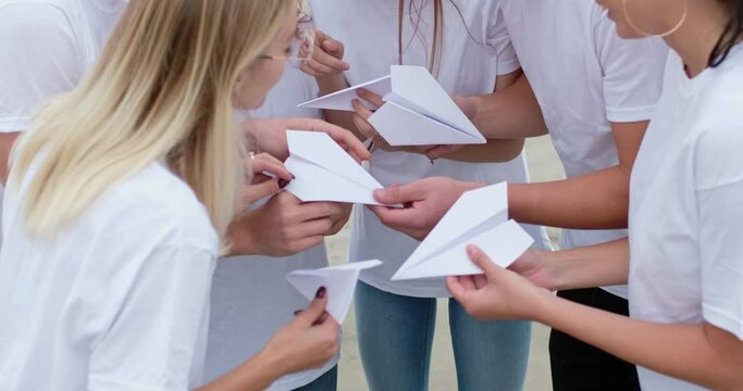 Team of young men and women wearing jeans and white t-shirts stand in circle raising hands with origami paper airplanes closeup