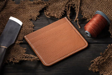 Making leather wallets. On a wooden table there is a leather wallet, a spool of thread and a knife.