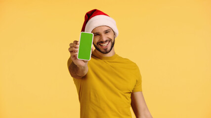 Fototapeta na wymiar pleased man in santa hat holding smartphone with green screen isolated on yellow