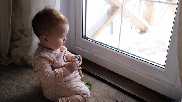 A cute plump Caucasian baby 10 months old is gnawing a toy on the floor near the door, window. Safe home space, real interior