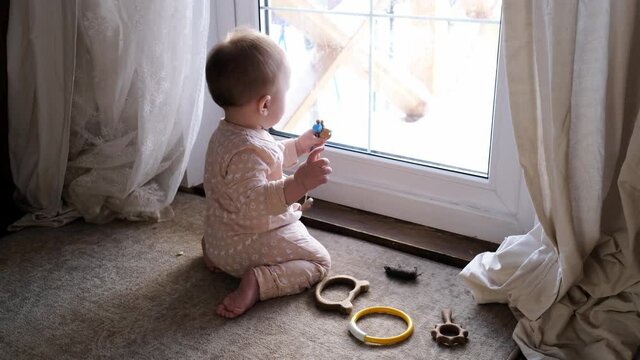 A cute plump Caucasian baby 10 months old is gnawing a toy on the floor near the door, window. Safe home space, real interior