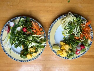 Close up of two plates of healthy vegan raw vegetable tofu and noodles meal with carrot cucumber coriander radish mango lime onion and chilli sauce flat lay plate view on wood surface interior