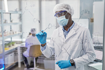 African chemist in protective wear writing chemical formulas on glass board, he examining new virus with his colleagues in the background
