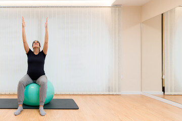 A pregnant woman doing pilates exercises sitting on a fitness ball with her arms stretched upward and looking up