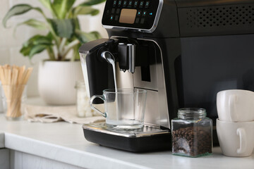 Modern coffee machine pouring milk into glass cup on white countertop in kitchen