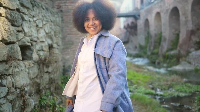 Fashion street style portrait of attractive young natural beauty African American woman with afro hair in blue coat posing outdoors. Happy tourist laughing walks through ancient sights fool around