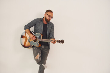 Standing in the studio. Man in casual clothes and with acoustic guitar is indoors