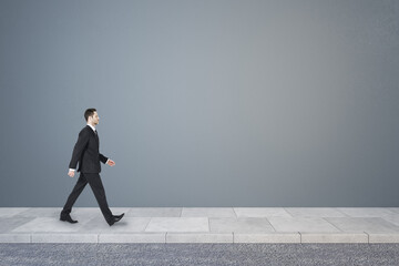 Side view of young business man walking in concrete interior with mock up place on wall. Success concept.