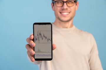 Portrait of positive brunette man holding cellphone and smiling, looking satisfied with device or mobile data tariffs. Indoor studio shot isolated on blue background