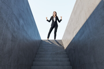 Happy european woman celebrating success on top of abstract concrete stairs with sunlight. Growth...