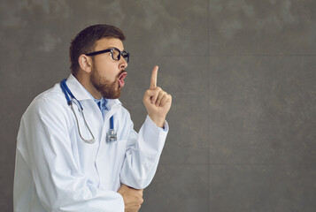 Young caucasian doctor surgeon pointing finger up looking inspired by genius thought showing good idea sign having clever solution in mind. Medicine and healthcare. Studio portrait shot over grey wall