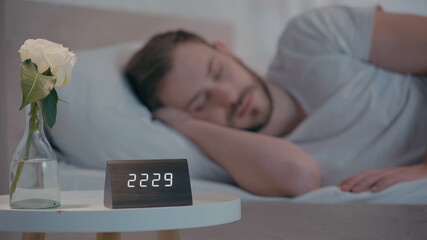 Rose and clock on bedside table near sleeping man at home.