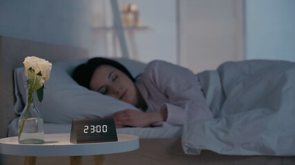 Flower and clock on bedside table near blurred brunette woman sleeping at home.