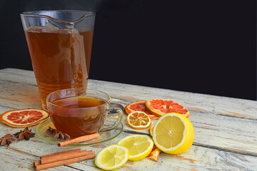 Glass cup of black tea with cinnamon sticks, anise star, lemon and dried fruit on white wooden table background. Jug of black tea. Large copy space
