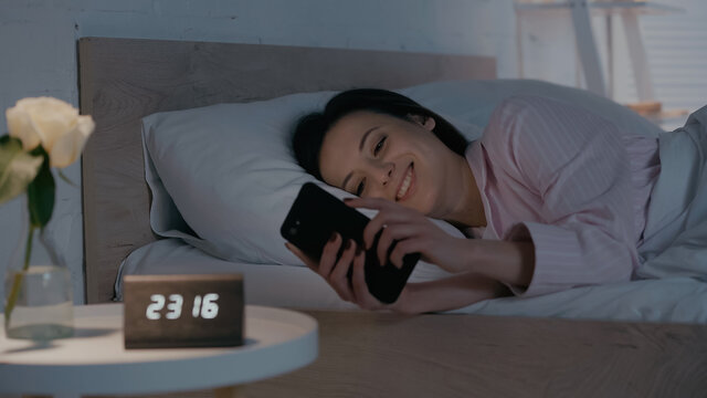 Cheerful woman using smartphone near blurred clock and plant in bedroom at night.