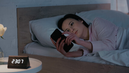 Brunette woman in pajamas using smartphone near blurred clock in bedroom.
