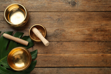 Golden singing bowls, mallets and monstera leaf on wooden table, flat lay. Space for text