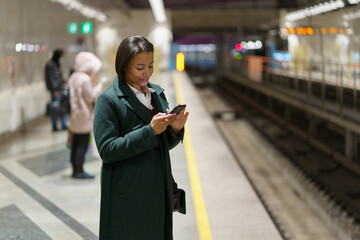 Selective focuse of young smiling african american woman at subway platform with smartphone...