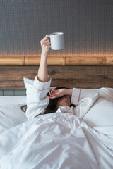 Hand of young woman lying in bed with coffee mug. Sleepy woman holding coffee cup relaxing in bed room. Female hand holding white Coffee Cup on bed in the morning. 