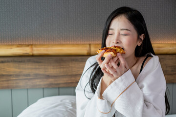 Happy young woman enjoy eating pizza on bed in hotel room. Beautiful female sitting in hotel bed...
