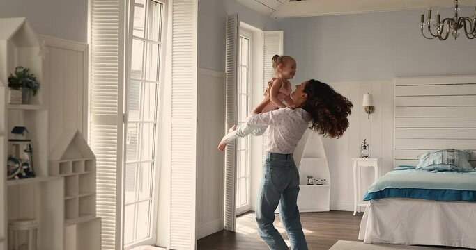 Lively mother holds her cute 4s daughter, throws up and whirling standing in modern bedroom at home. Family enjoy playtime and tender moments together. Happy motherhood, adoption, babysitting concept