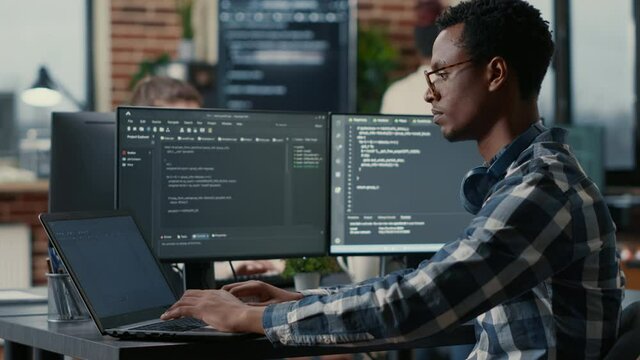 Portrait of african american developer using laptop to write code sitting at desk with multiple screens parsing algorithm in software agency. Coder working on user interface using portable computer.