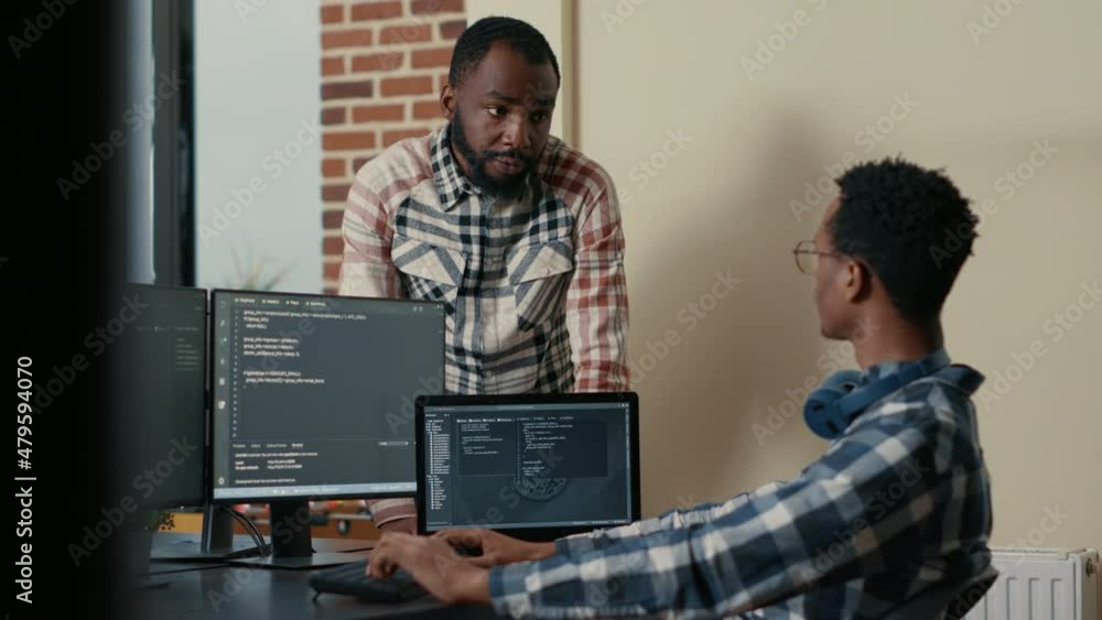 Poster software developer sitting at desk with multiple screens and laptop running code talking with collea